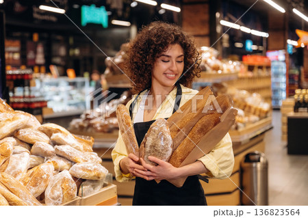 Standing in the supermarket and smiling. Female worker in apron is with bakery in hands Standing in the supermarket and smiling. Female worker in apron is with bakery in hands 136823564