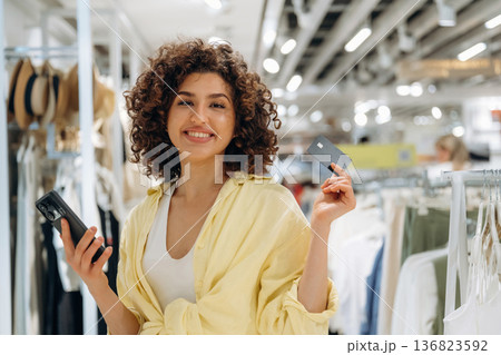 Curly hair, smile, credit card and smartphone. Woman is shopping in the clothing store 136823592