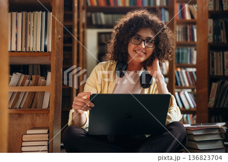 In headphones and with laptop, sitting. Beautiful woman is in the library 136823704