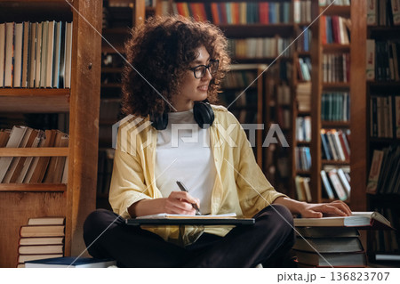 The writing process. With pen and headphones. Beautiful woman is in the library The writing process. With pen and headphones. Beautiful woman is in the library 136823707