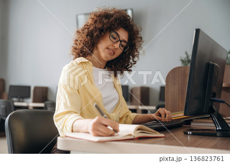 Making information research, with notepad and pen. Woman is in the library section with computers Making information research, with notepad and pen. Woman is in the library section with computers 136823761