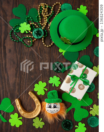 Top view flat lay of St Patrick's Day holiday with green gift, shamrocks, gold coins, lucky horseshoe, leprechaun hat on dark wooden background 136824309