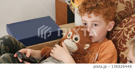 A cute boy with curly red hair is holding a stuffed monkey and looking sad 136826448