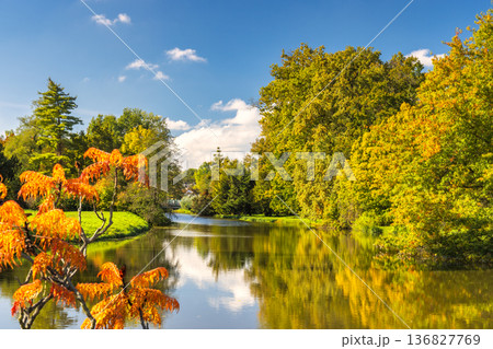Idyllic autumn scene features a tranquil river mirroring colorful trees under a clear blue sky with scattered clouds. Lednice Castle garden in South Moravia region of the Czech Republic, Europe. Idyllic autumn scene features a tranquil river mirroring colorful trees under a clear blue sky with scattered clouds. Lednice Castle garden in South Moravia region of the Czech Republic, Europe. 136827769
