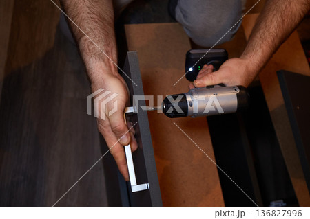 Person uses a power drill to assemble furniture in a room during daylight hours in a home setting Person uses a power drill to assemble furniture in a room during daylight hours in a home setting 136827996