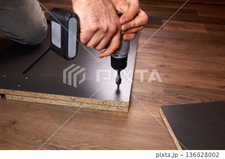 A person is installing a part of furniture by using a power drill. The setting shows wooden floors. A person is installing a part of furniture by using a power drill. The setting shows wooden floors. 136828002