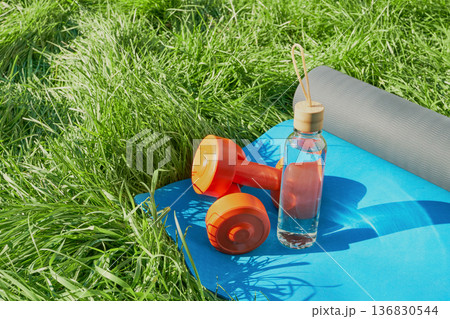 Glass bottle of water, dumbbells and blue yoga mat on green lawn Close-up Glass bottle of water, dumbbells and blue yoga mat on green lawn Close-up 136830544