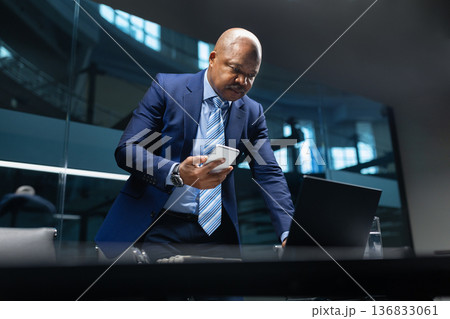 Mature black businessman standing at desk checking smartphone while working in office Mature black businessman standing at desk checking smartphone while working in office 136833061