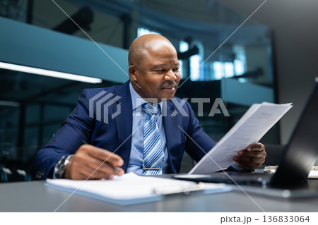 Smiling black businessman reading business report at desk in office, reviewing paperwork Smiling black businessman reading business report at desk in office, reviewing paperwork 136833064