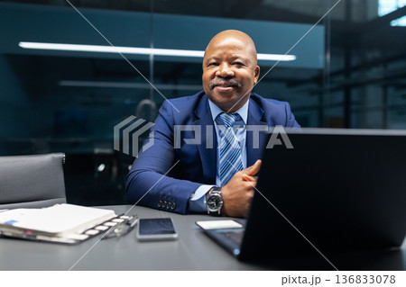 Black businessman smiling while sitting at desk with laptop in modern office 136833078