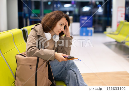 Stressed female traveler sitting in airport waiting area with headphones around neck Stressed female traveler sitting in airport waiting area with headphones around neck 136833102