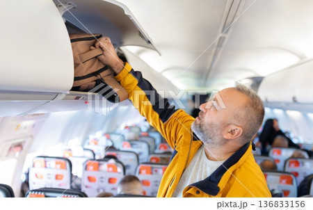 Adult caucasian male traveler placing backpack in airplane overhead compartment Adult caucasian male traveler placing backpack in airplane overhead compartment 136833156