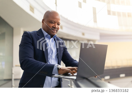 Focused mature black businessman working on laptop near office railing Focused mature black businessman working on laptop near office railing 136833182