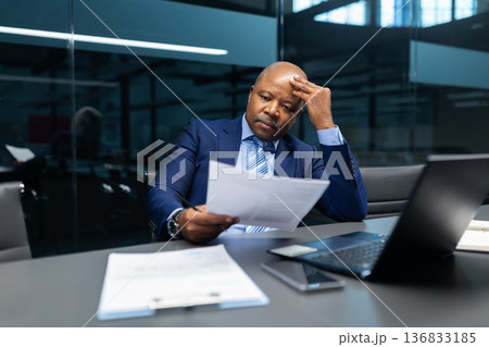 Mature black businessman reviewing printed document while touching forehead in modern office Mature black businessman reviewing printed document while touching forehead in modern office 136833185