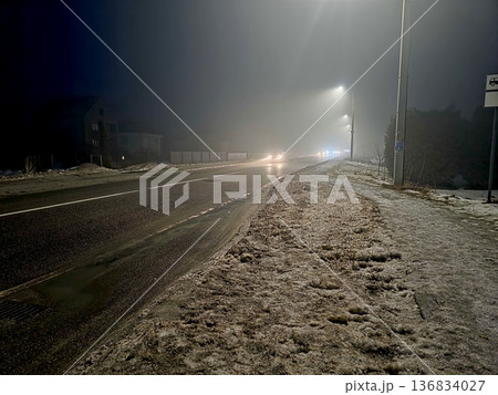 Night Road Covered in Snow With Distant Lights Shining Through Fog and Mist Night Road Covered in Snow With Distant Lights Shining Through Fog and Mist 136834027