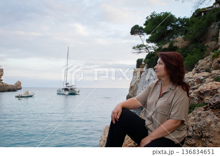 Middle aged woman sitting on the rock and enjoying sea views in Mallorca bay with yachts and boats. Calm seaside vacation in Spain. 136834651