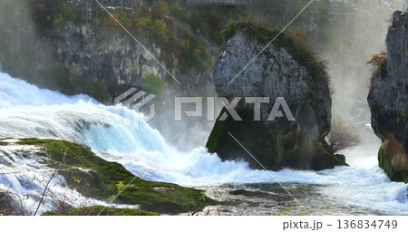 Water flows over rocks at a river during daytime in a natural setting with mist rising in the air near a forest 136834749