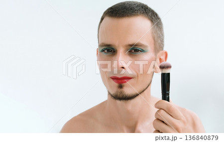 portrait of feminine happy gay man with makeup and cosmetics on face. Handsome adult transgender male with red lipstick on lips and eye shadows holds a blush brush on a white background 136840879