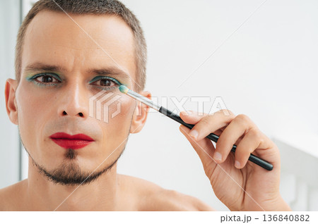 adult caucasian gay man with makeup and cosmetics on face. Transgender male with red lipstick applied eye shadow sparkle on his eyes with a brush on white background 136840882