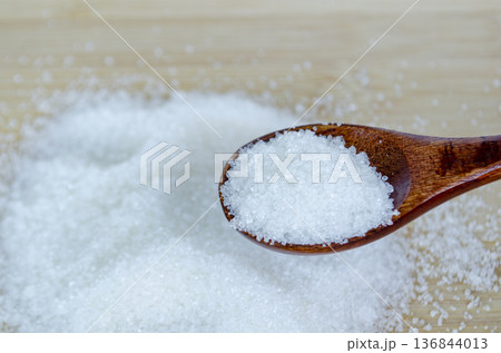 Granulated sugar scattered near wooden spoon representing excessive sugar intake and health risk awareness theme. Granulated sugar scattered near wooden spoon representing excessive sugar intake and health risk awareness theme. 136844013