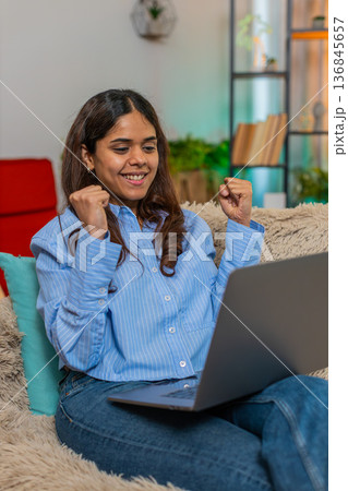 Indian woman celebrating good exam result on laptop with joyful jump and hand victory gesture home Indian woman celebrating good exam result on laptop with joyful jump and hand victory gesture home 136845657