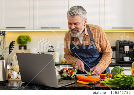 Middle-aged man preparing vegetable salad using laptop recipe in home kitchen for healthy diet meal Middle-aged man preparing vegetable salad using laptop recipe in home kitchen for healthy diet meal 136845789