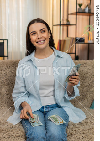 Young woman sitting on sofa thoughtfully counting cash dollars budgeting money sighing setting aside Young woman sitting on sofa thoughtfully counting cash dollars budgeting money sighing setting aside 136845796