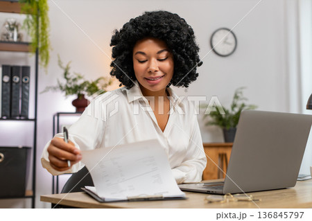 African American businesswoman reviews paperwork nods satisfied seeing laptop data confirm records African American businesswoman reviews paperwork nods satisfied seeing laptop data confirm records 136845797