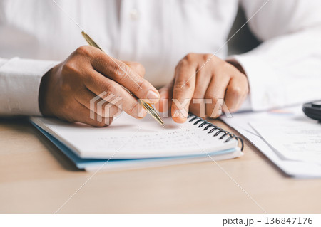 Hand writing notes in a notebook beside a calculator on office desk for budgeting and accounting. Personal finance planning, expense tracking, tax calculation, soft focus background, copy space. Hand writing notes in a notebook beside a calculator on office desk for budgeting and accounting. Personal finance planning, expense tracking, tax calculation, soft focus background, copy space. 136847176