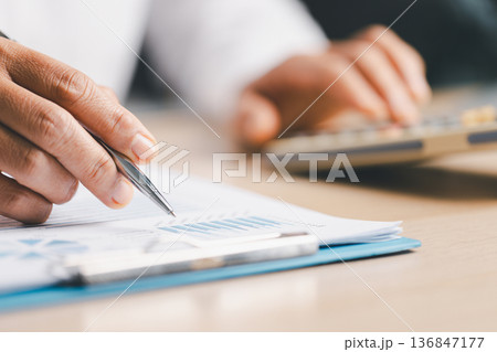 Hand writing notes in a notebook beside a calculator on office desk for budgeting and accounting. Personal finance planning, expense tracking, tax calculation, soft focus background, copy space. 136847177