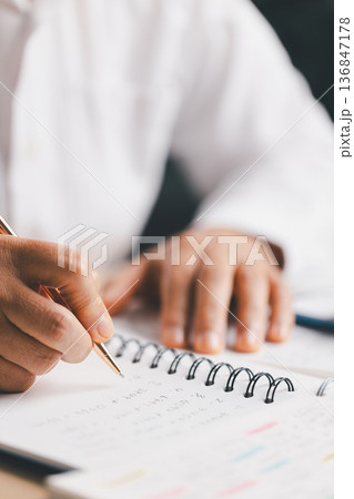 Hand writing notes in a notebook beside a calculator on office desk for budgeting and accounting. Personal finance planning, expense tracking, tax calculation, soft focus background, copy space. Hand writing notes in a notebook beside a calculator on office desk for budgeting and accounting. Personal finance planning, expense tracking, tax calculation, soft focus background, copy space. 136847178