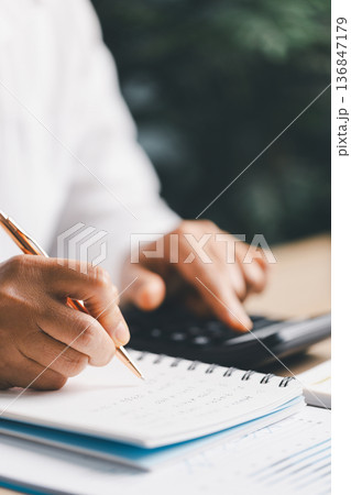 Hand writing notes in a notebook beside a calculator on office desk for budgeting and accounting. Personal finance planning, expense tracking, tax calculation, soft focus background, copy space. Hand writing notes in a notebook beside a calculator on office desk for budgeting and accounting. Personal finance planning, expense tracking, tax calculation, soft focus background, copy space. 136847179