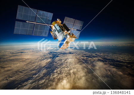 Detailed view of a satellite in space, featuring metallic structure, antennas, and large solar panels, orbiting above Earth's blue atmosphere and orange-tinted cloud layers against the dark void. 136848141