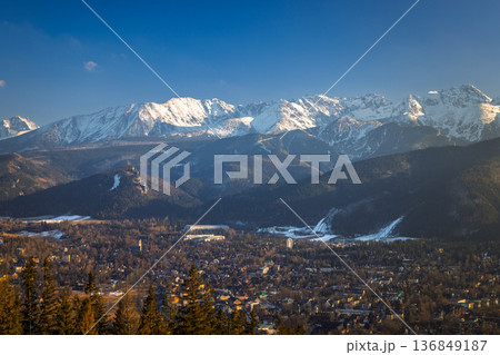 Majestic snow-capped mountains tower over a sprawling town nestled in a valley, beneath a clear blue winter sky. Zakopane town at the foot of the Tatra Mountains in Poland, Europe. Majestic snow-capped mountains tower over a sprawling town nestled in a valley, beneath a clear blue winter sky. Zakopane town at the foot of the Tatra Mountains in Poland, Europe. 136849187