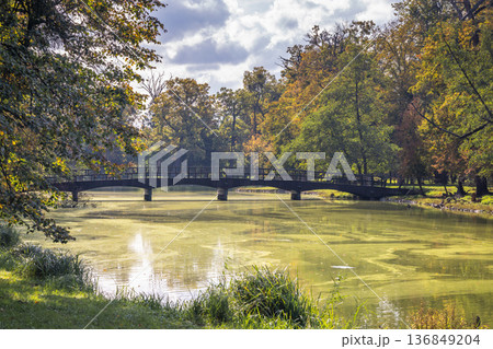 An old wooden bridge spans across a tranquil body of water, surrounded by trees changing color during fall. Lednice Castle garden in South Moravia region of the Czech Republic, Europe. An old wooden bridge spans across a tranquil body of water, surrounded by trees changing color during fall. Lednice Castle garden in South Moravia region of the Czech Republic, Europe. 136849204