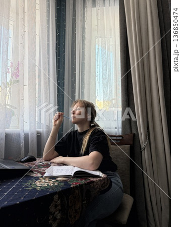 Thoughtful woman sitting at a table over her notes with a pen at her chin and looking out the window 136850474