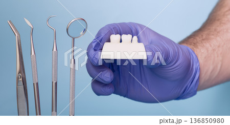 Dentist Hand in Blue Medical Glove Holding Dental Crown Shade or Prosthetic Tooth Model with Dental Instruments on Light Blue Background 136850980