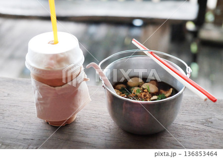 Thai boat noodles with chicken soup and pork balls in aluminum bowl with iced coffee in plastic glass on wooden table Thai boat noodles with chicken soup and pork balls in aluminum bowl with iced coffee in plastic glass on wooden table 136853464