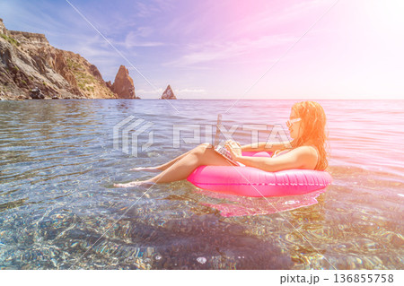 Remote work digital nomad freelance woman sitting on pink inflatable ring using laptop while floating in clear blue ocean water under sun flare Remote work digital nomad freelance woman sitting on pink inflatable ring using laptop while floating in clear blue ocean water under sun flare 136855758