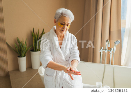 Mature adult old woman applying moisturizing body lotion on hands. Senior lady rubbing nourishing anti age cream doing morning daily skincare routine in bathroom. Beauty treatment spa body skin care 136856181