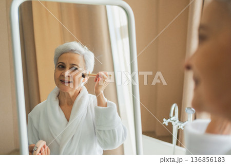 Mature adult old woman with smooth soft perfect skin applying rouge with brush on cheek looking at mirror doing daily beauty make up routine. Senior woman putting blush on her face. Anti age makeup 136856183