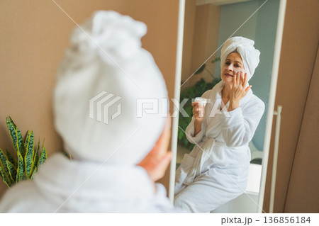 Mature adult old woman looking at mirror applying nourishing anti age face cream doing daily beauty routine. Senior woman with white mask creme on skin gets facial treatment Skin care aging skincare Mature adult old woman looking at mirror applying nourishing anti age face cream doing daily beauty routine. Senior woman with white mask creme on skin gets facial treatment Skin care aging skincare 136856184