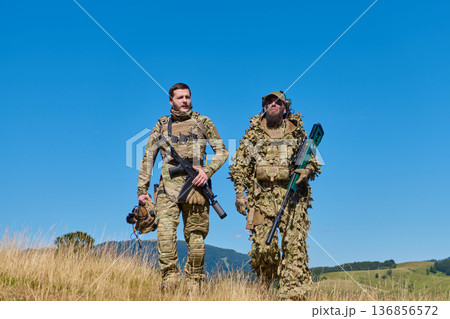 Two Soldiers in Camouflage and Tactical Gear Walking Through Open Field With Rifles 136856572