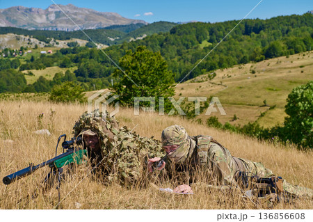 Military Snipers in Camouflage Ghillie Suits Lying in a Grass Field on a Mountain Hillside Military Snipers in Camouflage Ghillie Suits Lying in a Grass Field on a Mountain Hillside 136856608