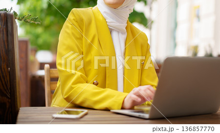 Woman works on laptop at outdoor cafe while wearing a yellow jacket and a hijab Woman works on laptop at outdoor cafe while wearing a yellow jacket and a hijab 136857770