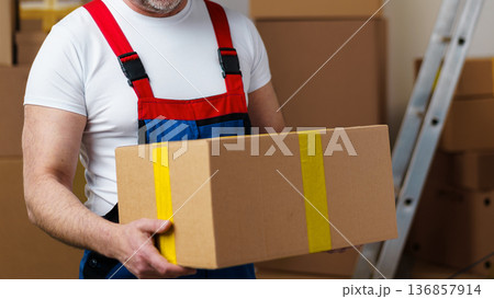 Worker carries a large cardboard box in a warehouse during the day 136857914