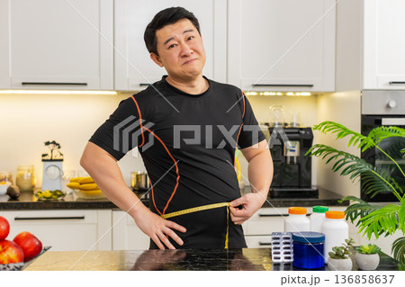 Heavy Asian adult man measures waist with tape upset about big belly fat weight gain at home kitchen Heavy Asian adult man measures waist with tape upset about big belly fat weight gain at home kitchen 136858637