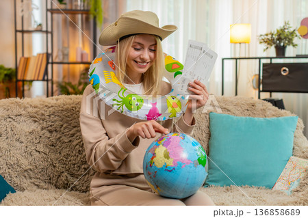 Woman sitting on sofa at home planning trip holding tickets marking route on globe with dreamy smile Woman sitting on sofa at home planning trip holding tickets marking route on globe with dreamy smile 136858689