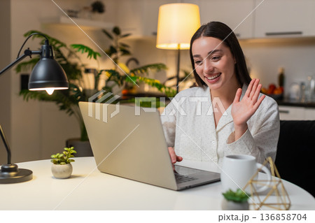 Young woman on laptop webcam waves hello speaks shows charts to client listens nods online evening Young woman on laptop webcam waves hello speaks shows charts to client listens nods online evening 136858704