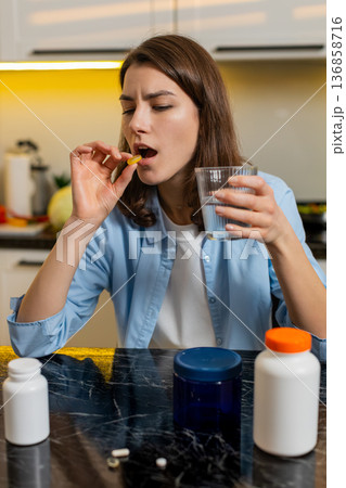 Young woman feeling sick taking medicine at home kitchen with water glass for illness treatment care Young woman feeling sick taking medicine at home kitchen with water glass for illness treatment care 136858716